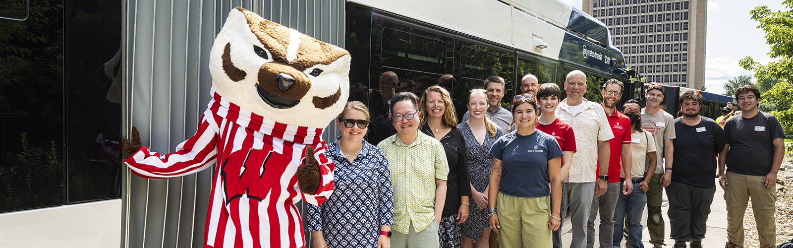 A person dressed in the Bucky Badger mascot with about ten people standing in front of a Madison metro bus